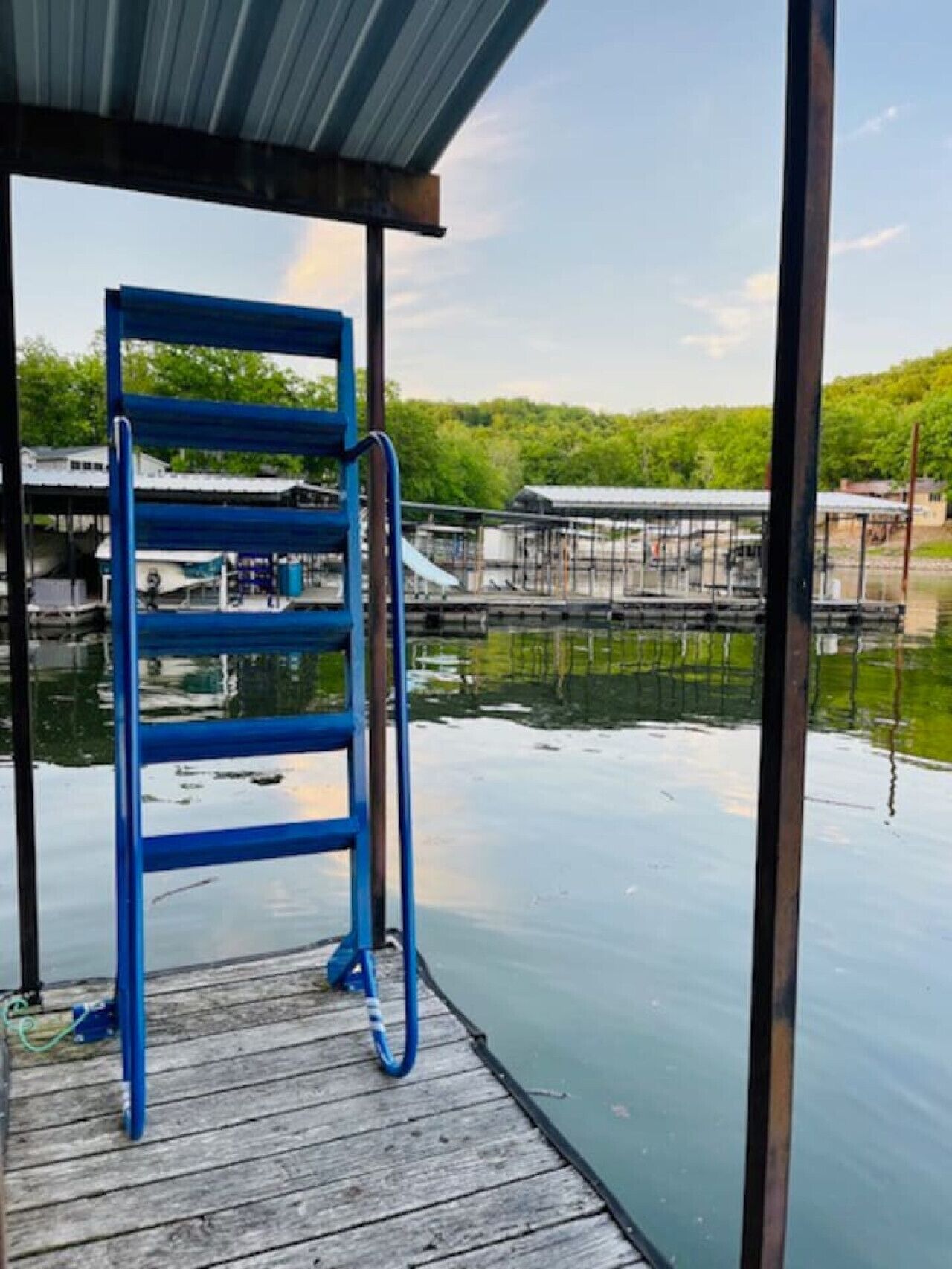 Een blauwe metalen ladder op een houten steiger die boven kalm water uitsteekt, met een overdekte ruimte erboven en een boot afgemeerd op de achtergrond.
