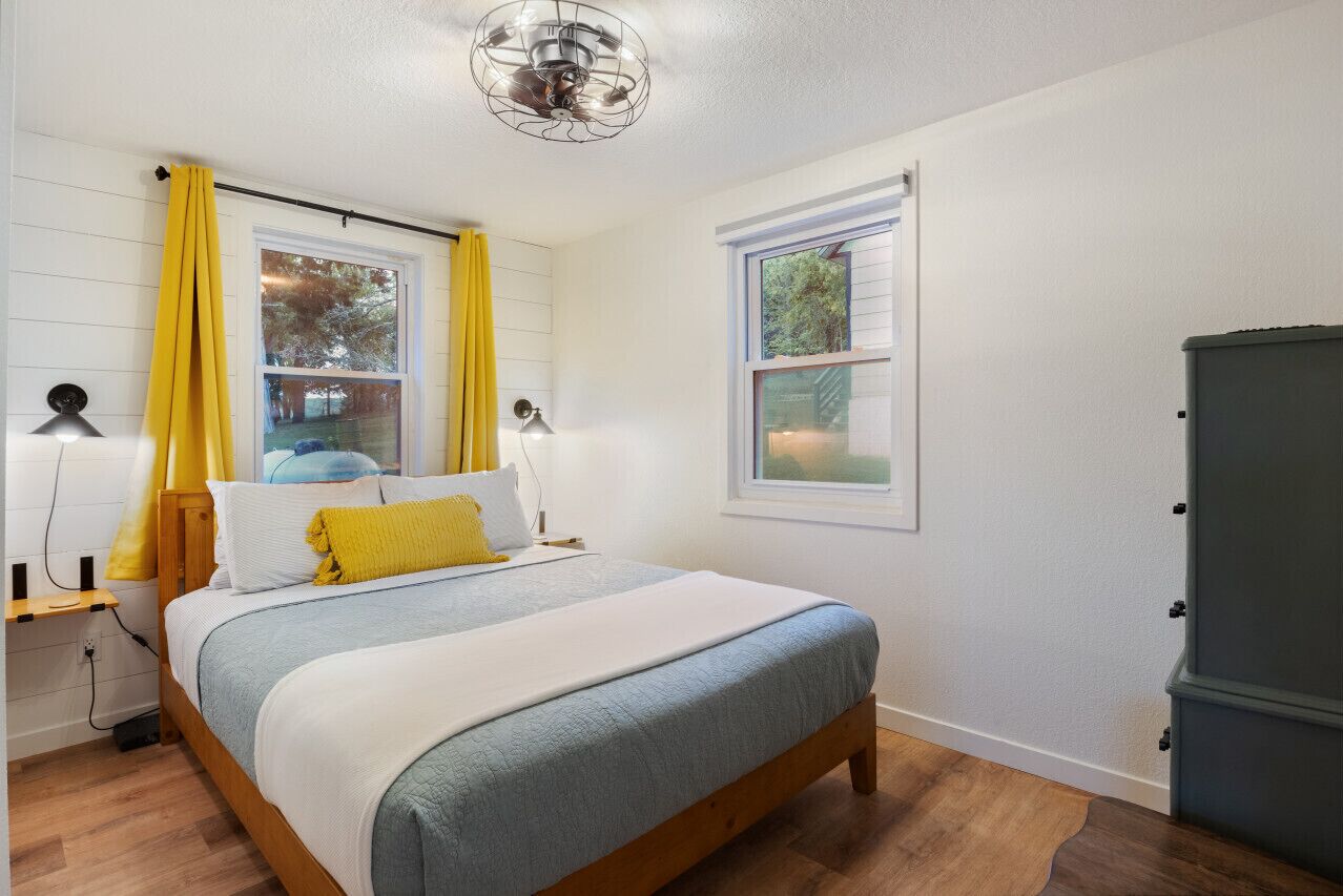 Ein Schlafzimmer mit einem Bett, zwei Nachttischlampen, einem Fenster mit Vorhängen, einem Deckenventilator und einem Kleiderschrank.