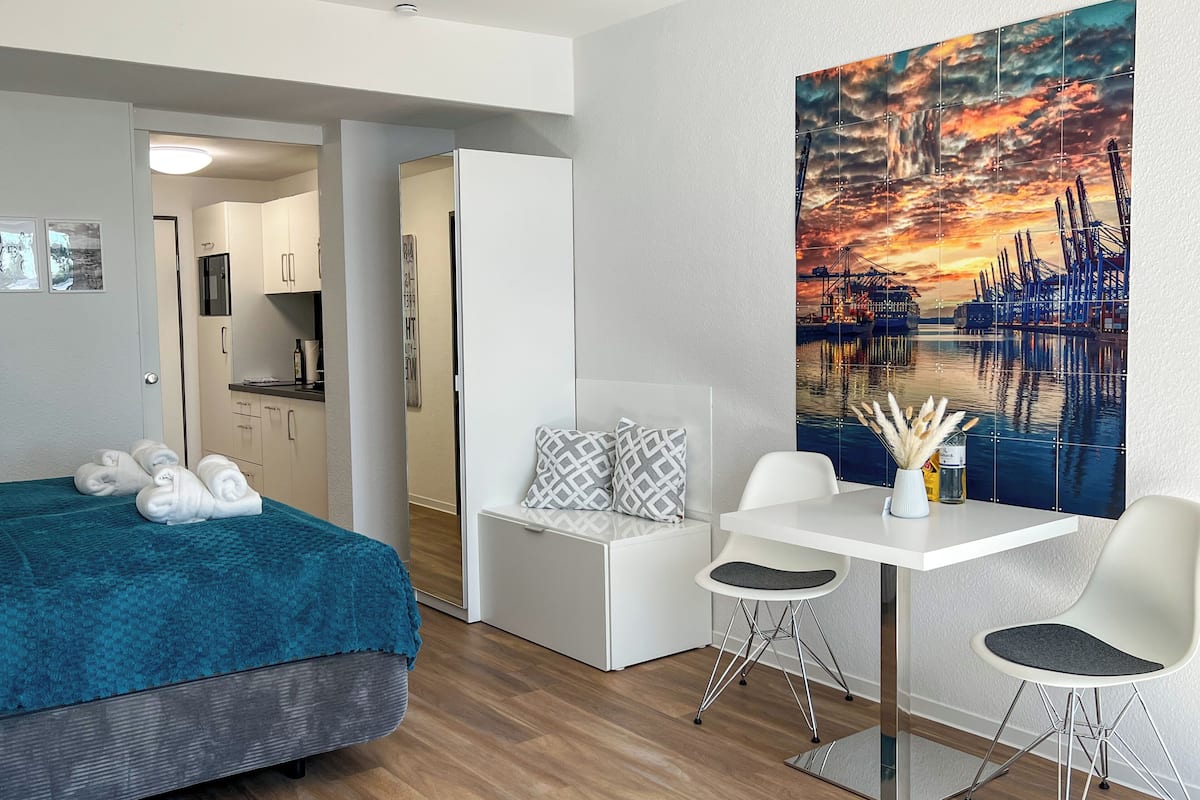 A modern hotel room with a bed, a desk, and a large painting of a sunset over a harbor.