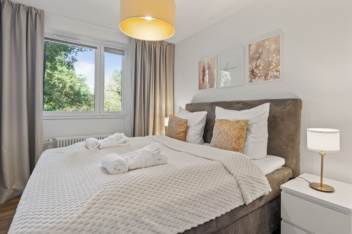 A neatly made bed with a white quilt, a bedside lamp, and a window with curtains.