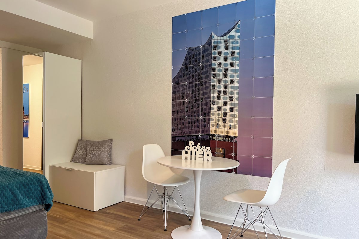 A modern hotel room with a wooden floor, a white sofa, a round table with two chairs, and a large cityscape painting on the wall.