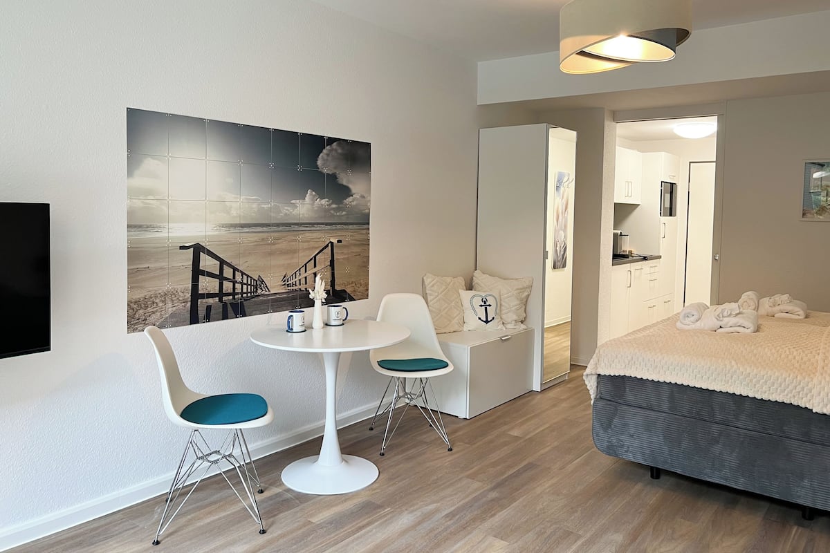 A hotel room with a bed, a desk with two chairs, a painting of a beach, and a modern lamp.