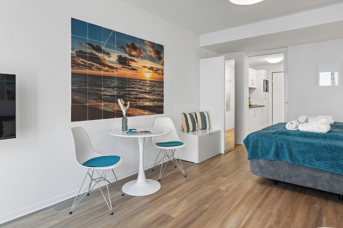A modern hotel room with a wooden floor, a bed with a teal blanket, a white desk with two chairs, a large sunset painting on the wall, and a TV mounted on the wall.