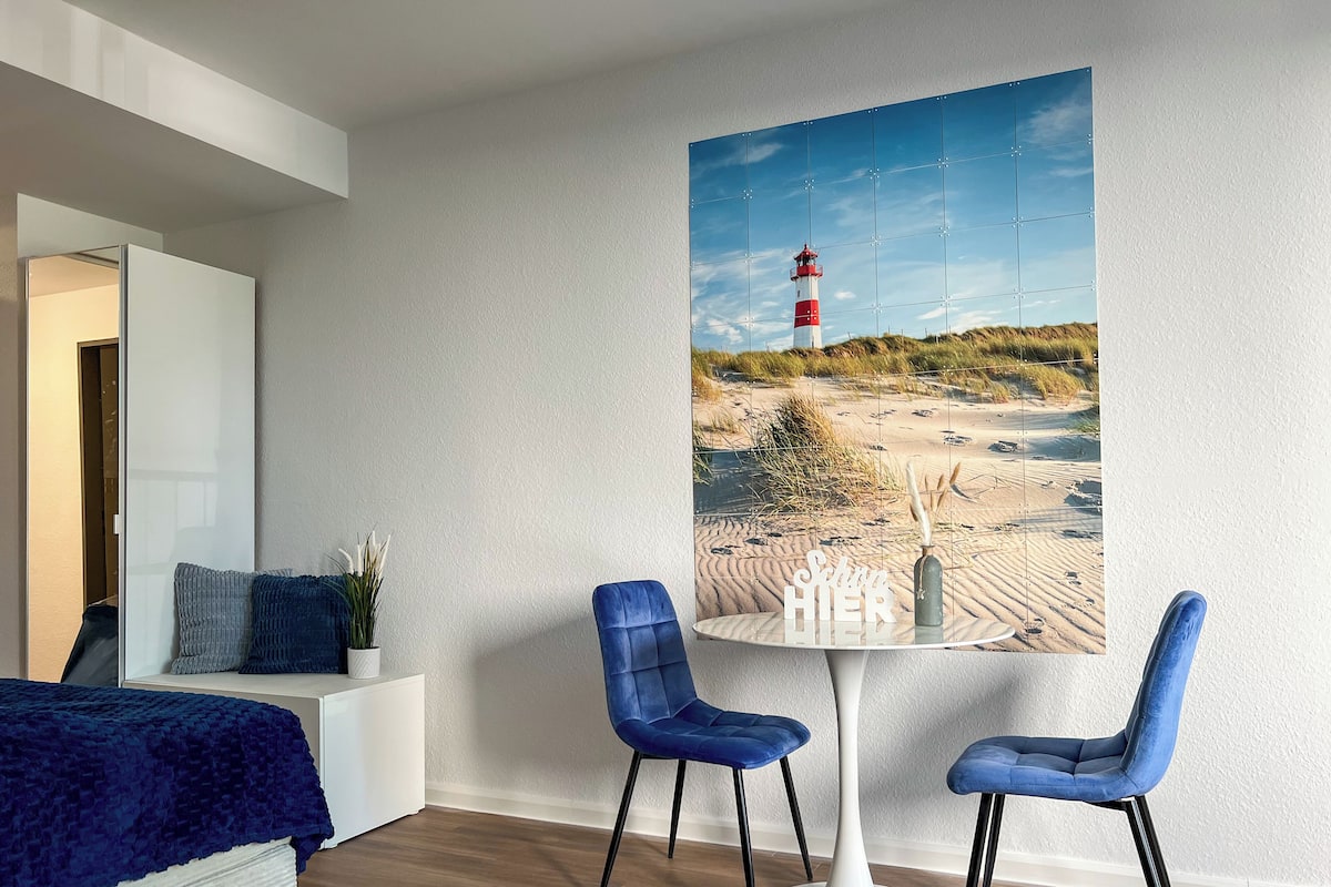 A modern room with a white bed, a blue sofa, and a round table with two blue chairs. A large wall-mounted picture of a lighthouse and beach is visible.