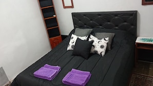 A neatly made bed with a black headboard, two purple towels, and patterned pillows. A wooden shelf and a mirror are on the wall beside the bed. - LODGING NAOMI VILLA UNIÓN PROVINCE OF LA RIOJA  (Villa Unión)