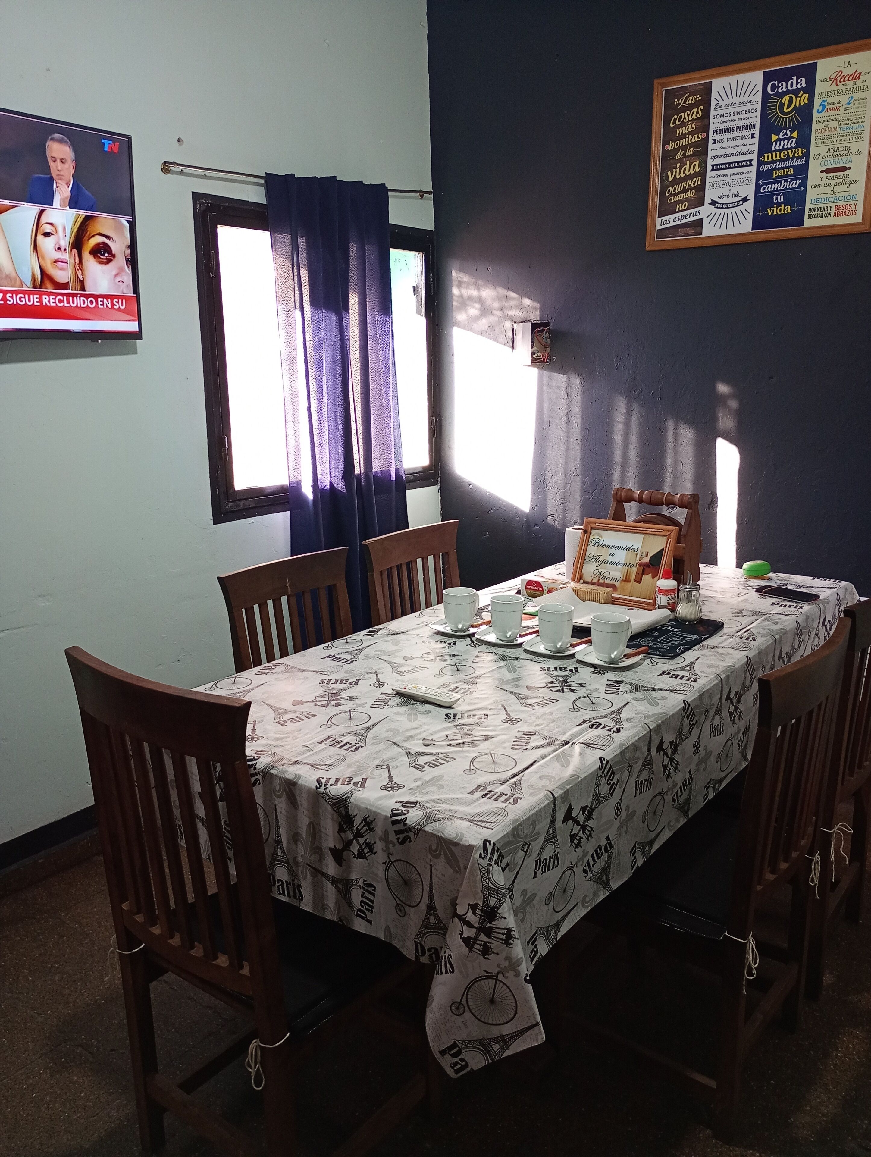 Un comedor con una mesa dispuesta para cuatro, sillas, una ventana con cortinas y un cartel en la pared.