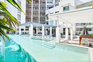 Una piscina con sillas de descanso y sombrillas, rodeada de edificios blancos y palmeras.