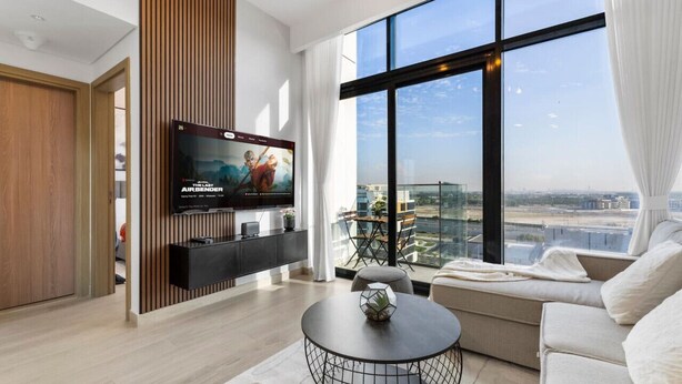 Una sala de estar moderna con un televisor grande, un sofá, una mesa redonda de centro y vistas a la ciudad a través de amplios ventanales.