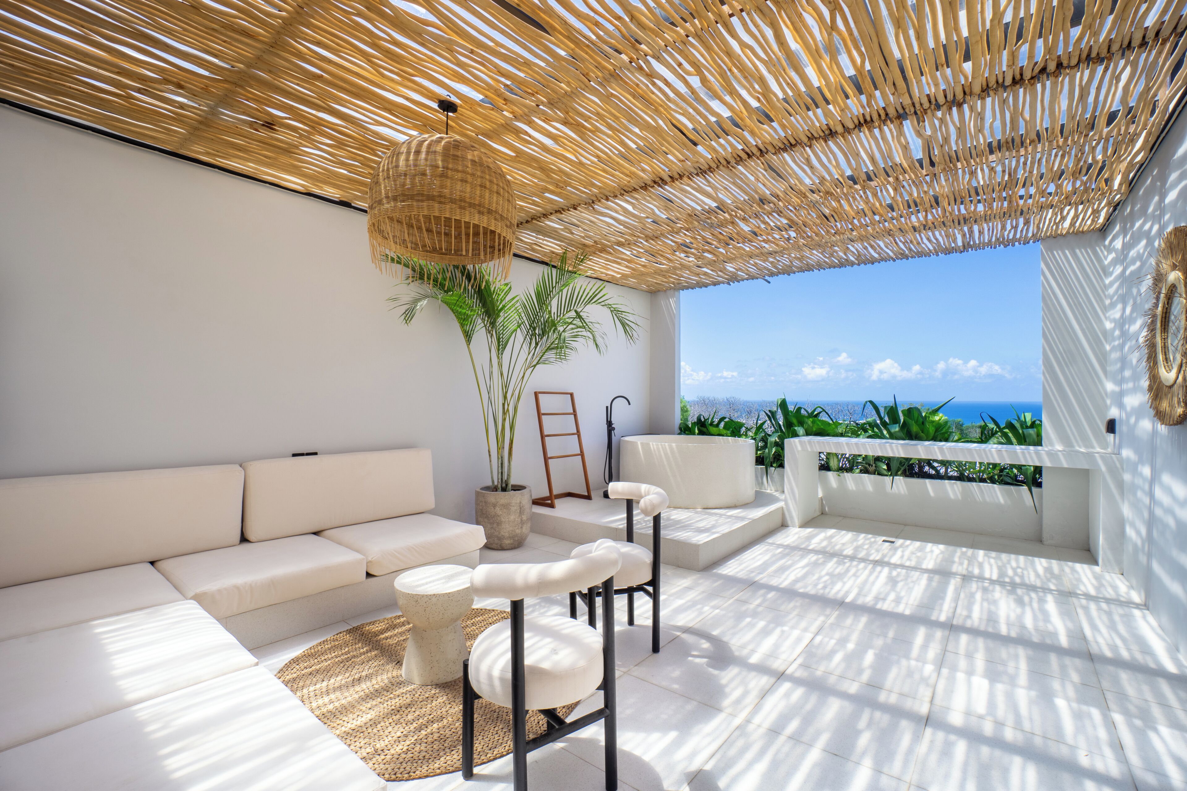 A sunlit outdoor seating area with a white sofa, two chairs, a woven ceiling, and a view of the ocean.