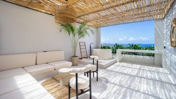 A sunlit outdoor seating area with a white sofa, two chairs, a woven ceiling, and a view of the ocean.