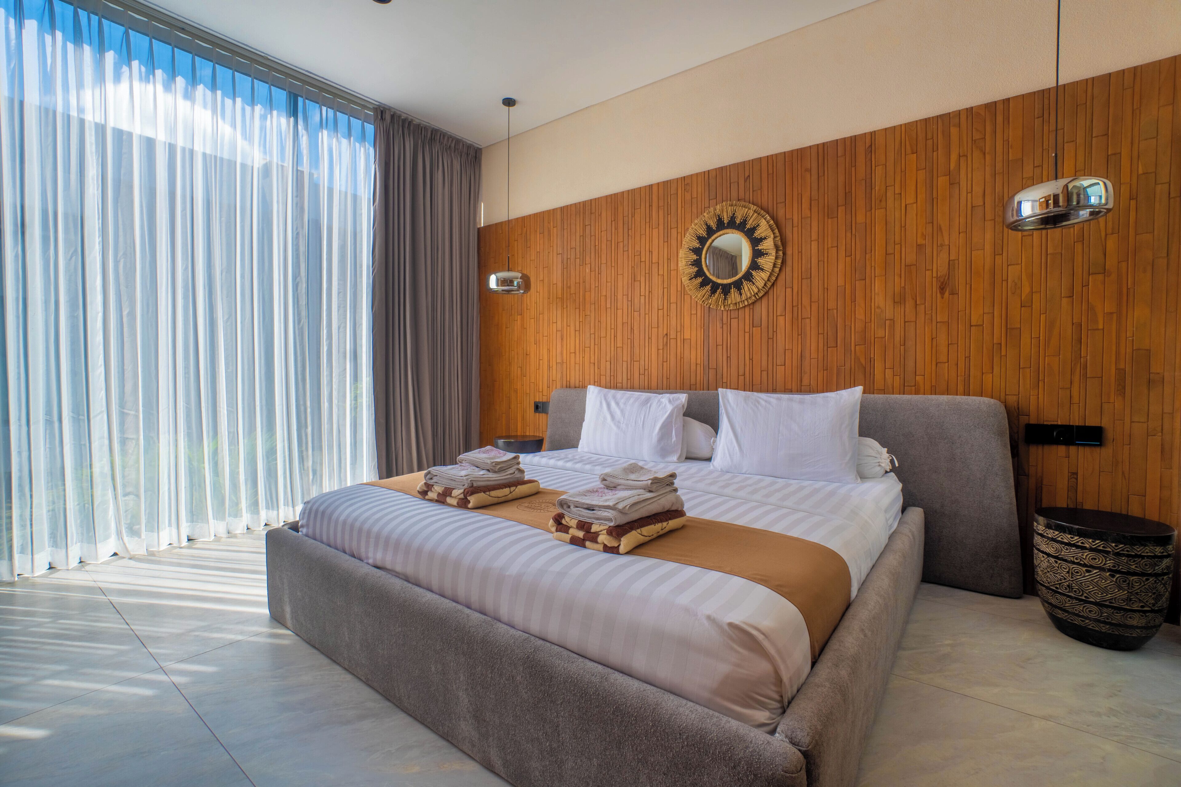 Un dormitorio moderno con una cama grande, pared de paneles de madera y vistas a través de cortinas translúcidas.