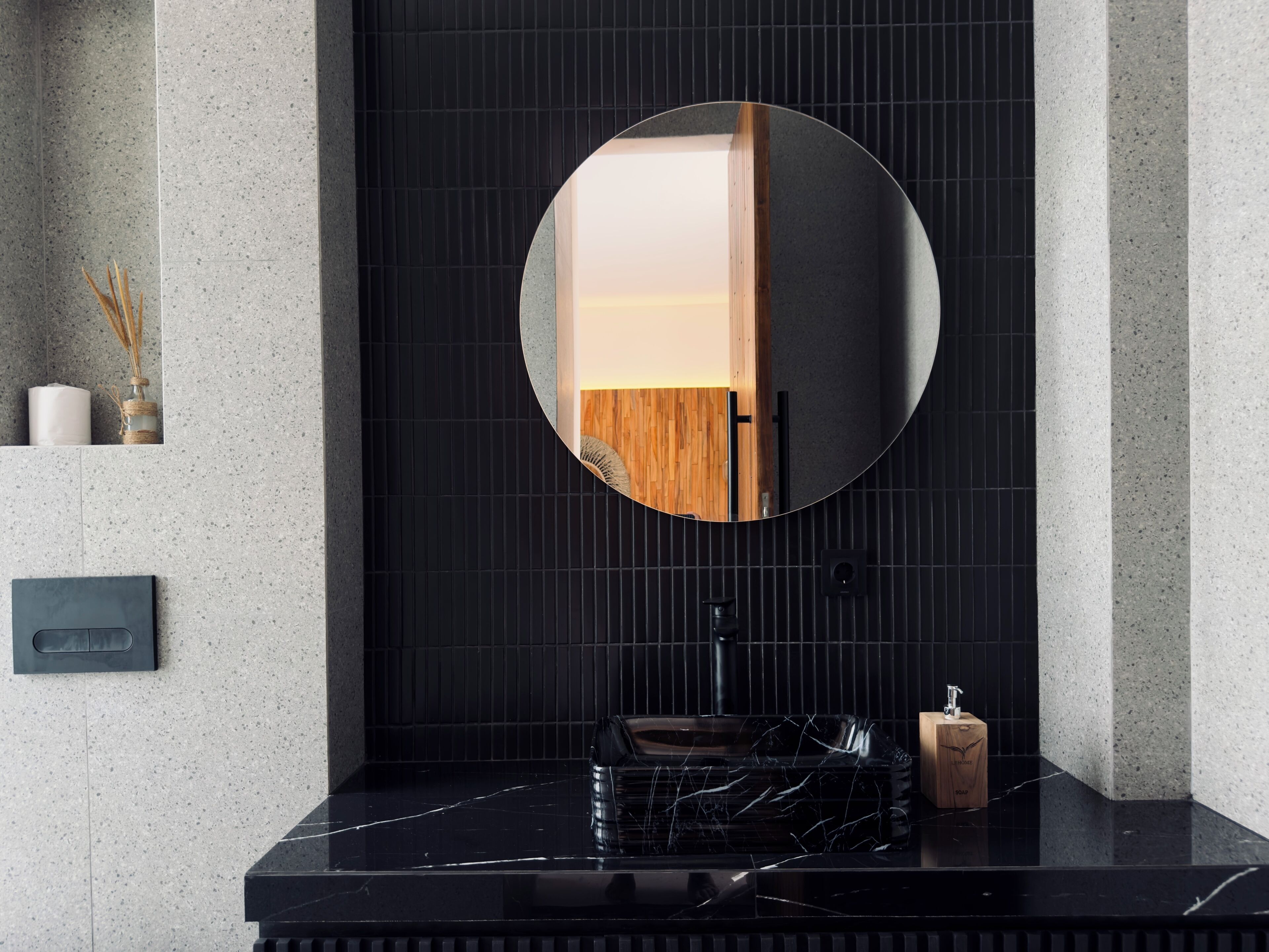 A modern bathroom with a black marble countertop, a round mirror, and a wooden door.