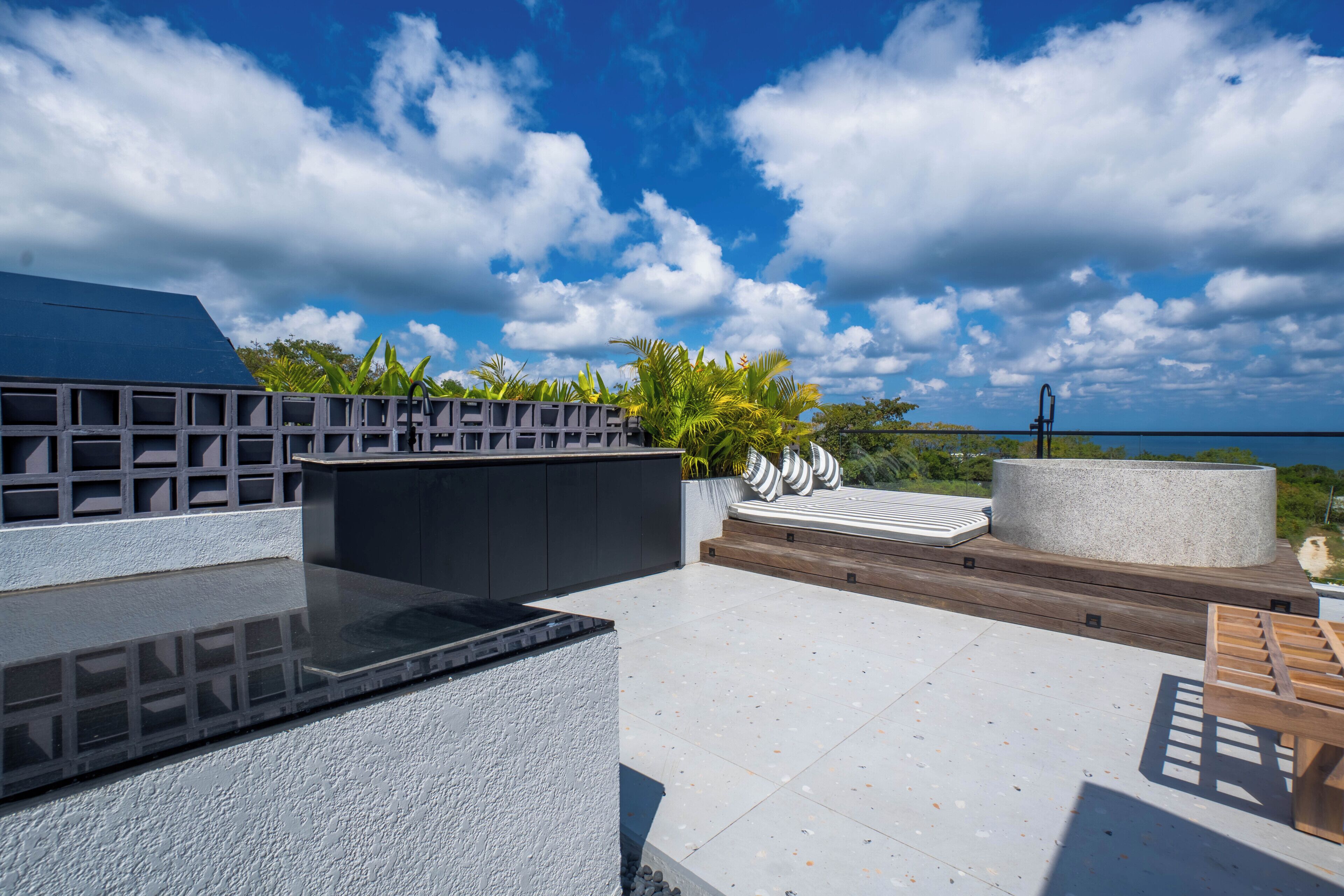 En takterrass med modern design, med en sittgrupp bestående av kuddar och ett litet bord, omgiven av grönska och en klar himmel.