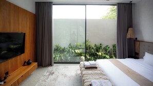 A modern bedroom with a large TV, a bed, a bench, and a view of greenery through the sliding glass door. - Rahya Villas Complex (Kutuh)