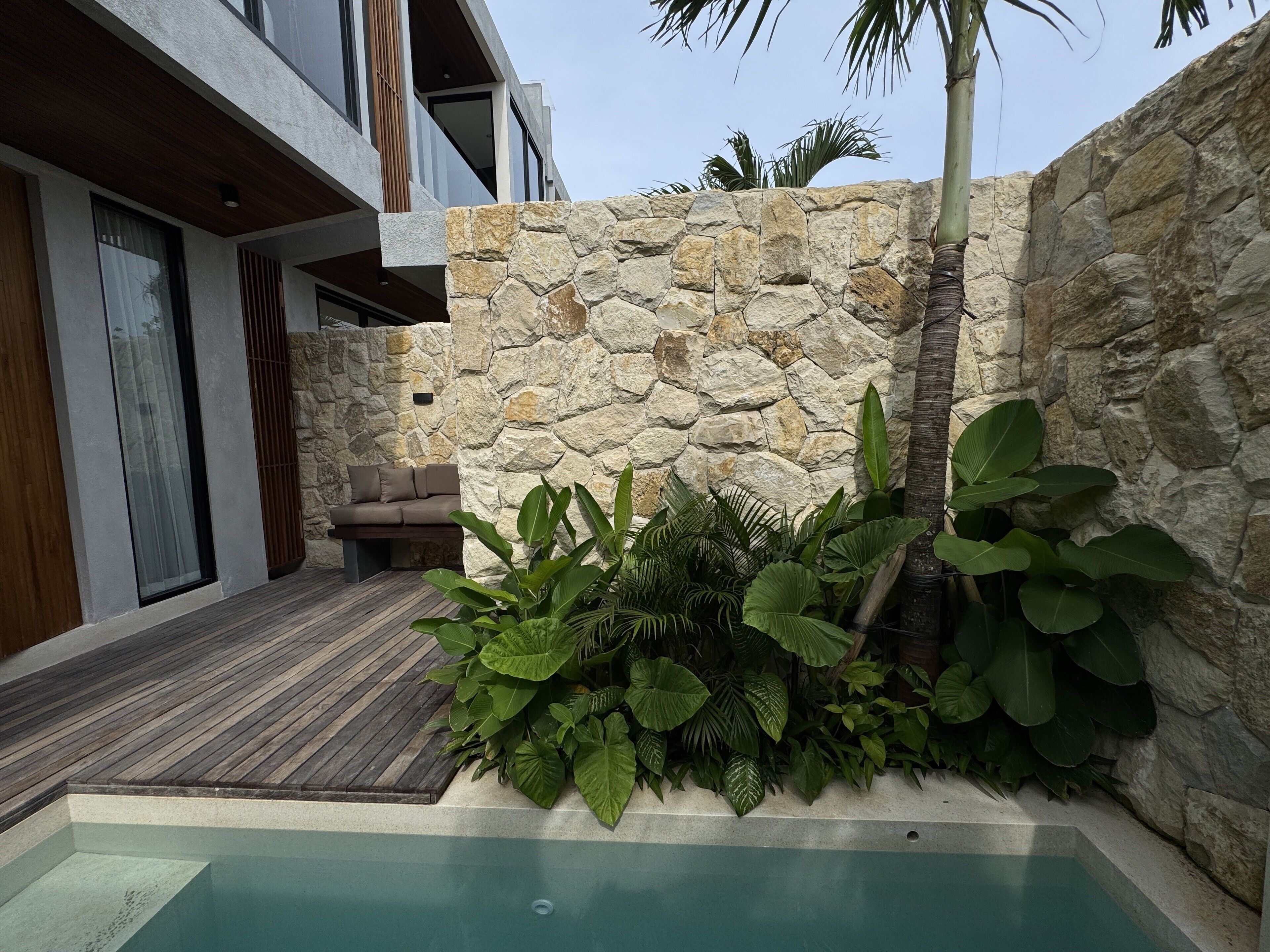 A modern outdoor pool area with a stone wall, wooden deck, and tropical plants.