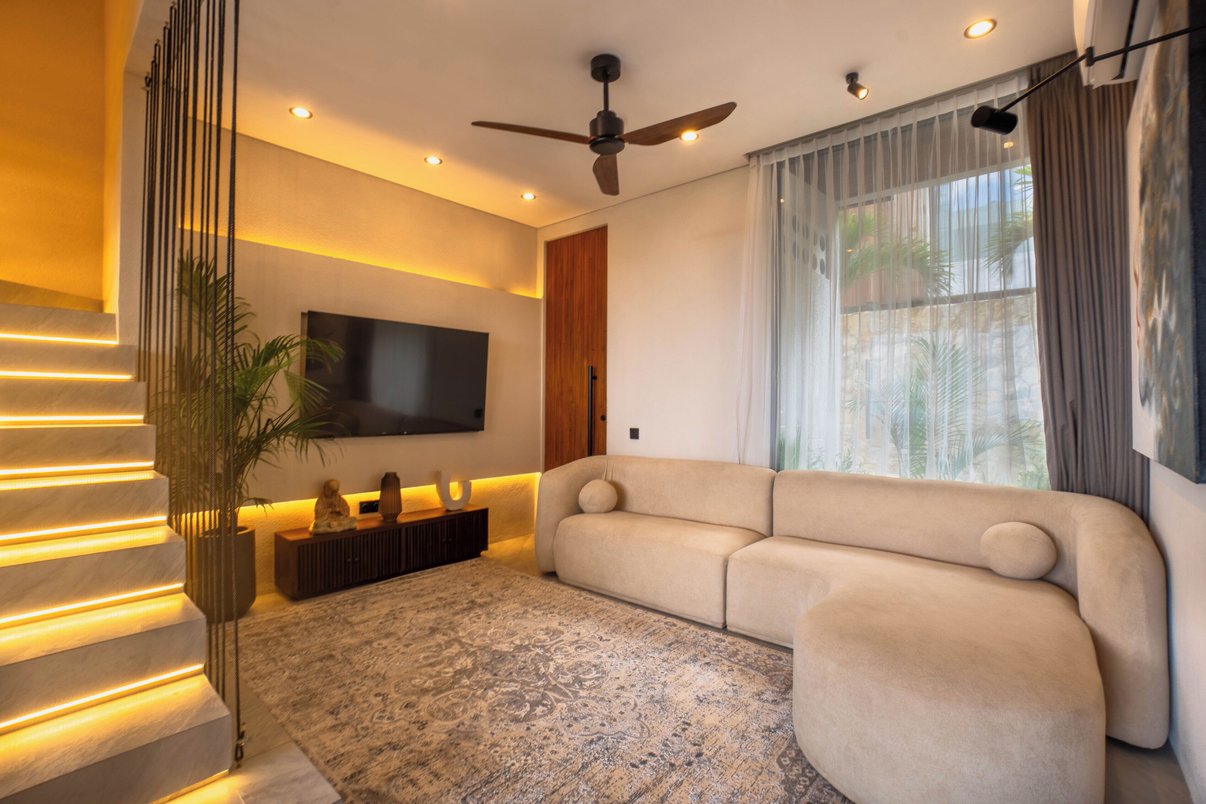 Een moderne woonkamer met een hoekbank, een flatscreen-televisie aan de muur, een plafondventilator en een groot raam met gordijnen.
