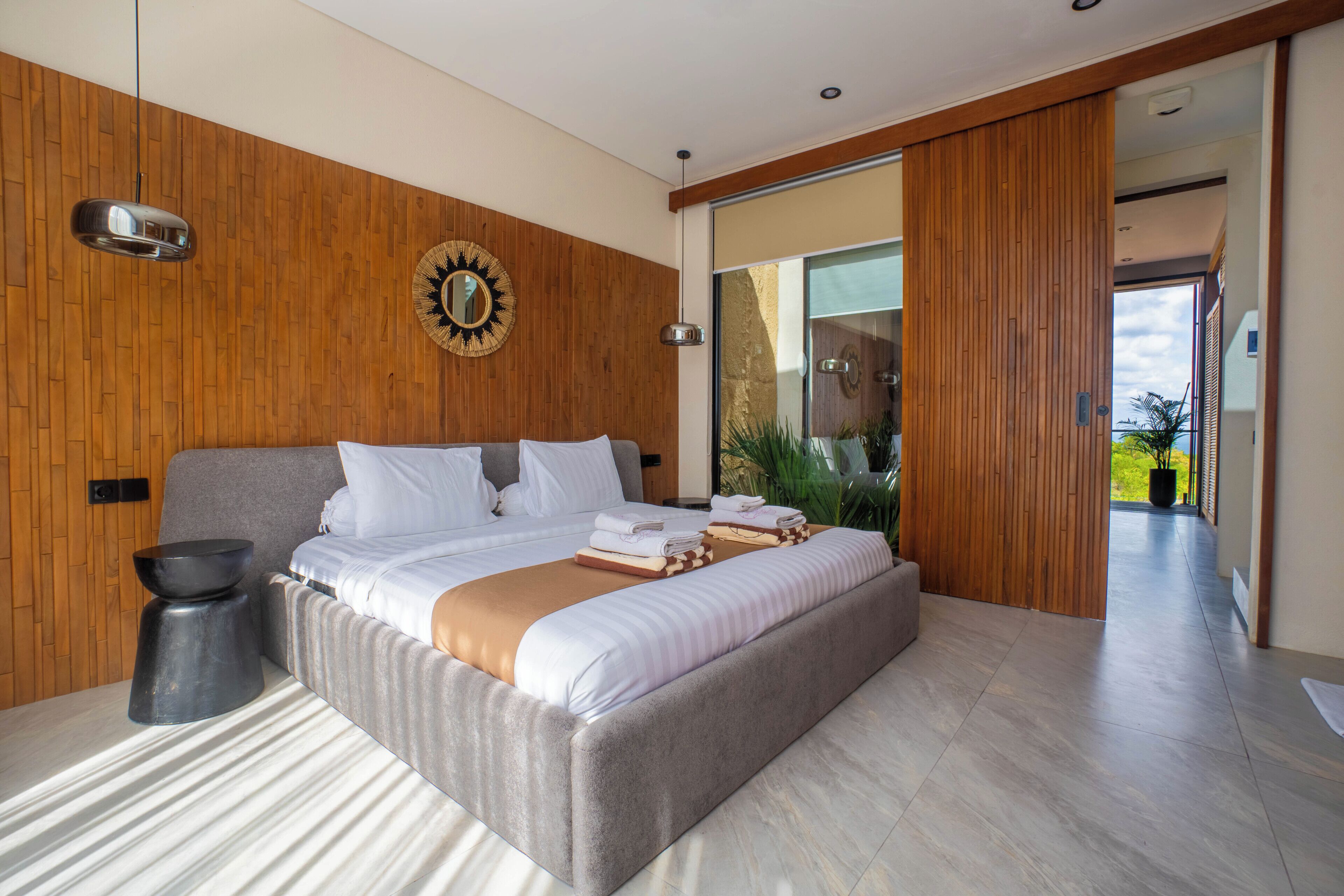 Ein modernes Schlafzimmer mit einem großen Bett, Holzpaneelen, einem runden Spiegel und Blick auf Grünflächen.