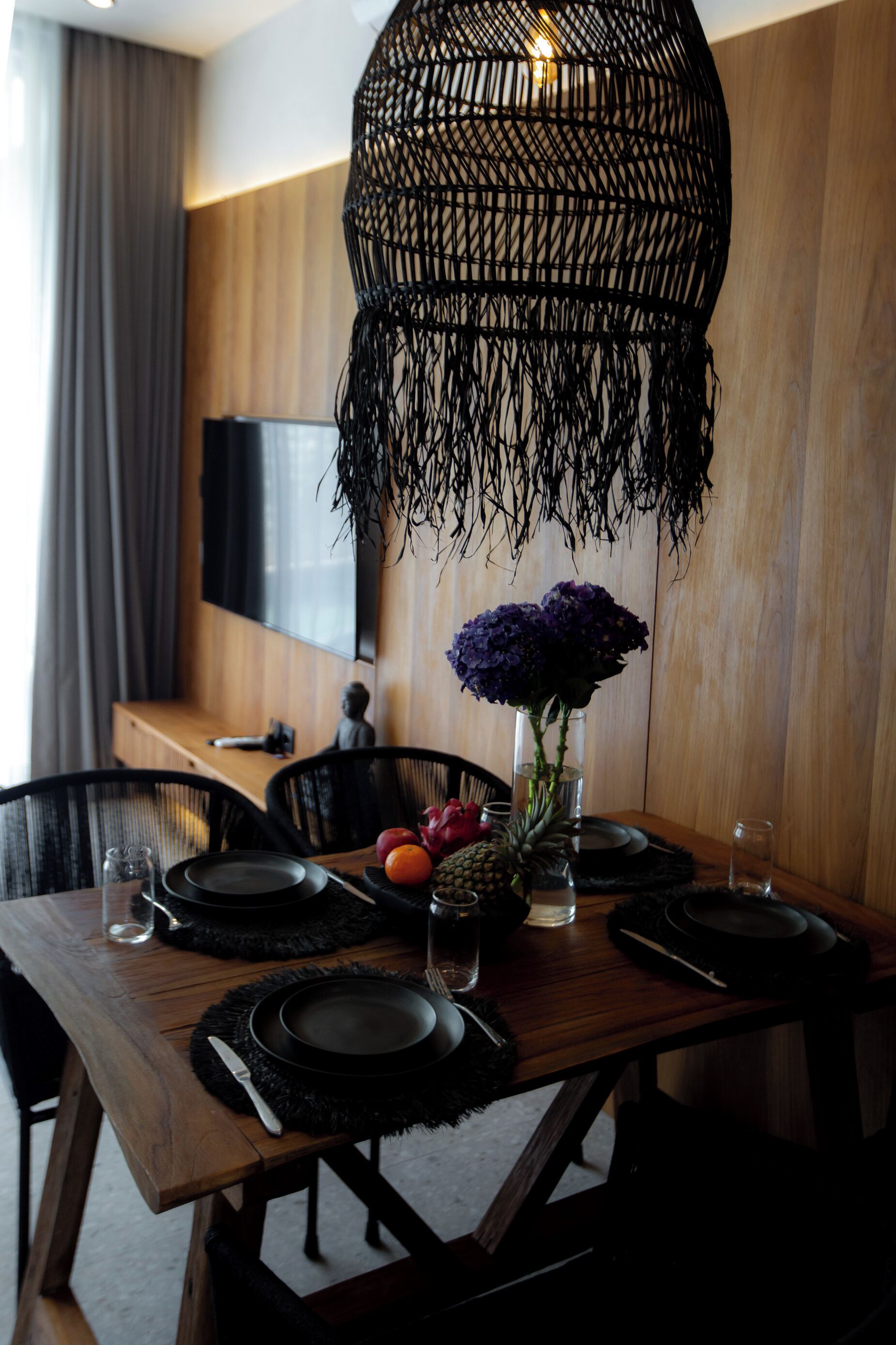 Mesa de comedor dispuesta con platos negros, cubiertos y un jarrón con flores moradas.