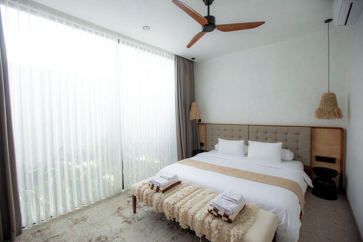 Une chambre d’hôtel avec un grand lit, un ventilateur de plafond, une fenêtre avec des rideaux, une lampe de chevet et un petit tabouret avec des serviettes.