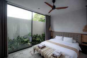 A modern bedroom with a large bed, a ceiling fan, and a view of greenery through a sliding door.