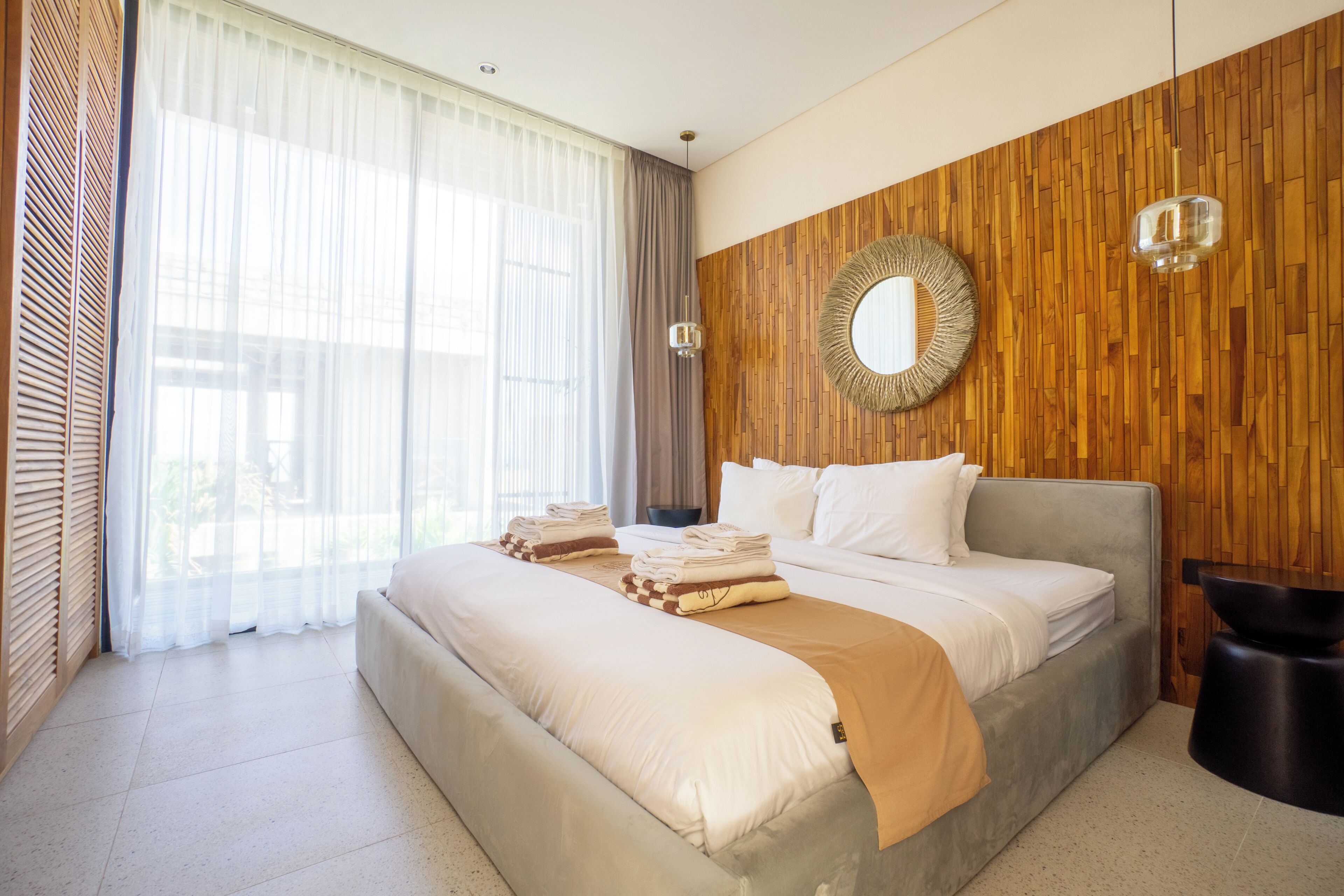 Une chambre moderne avec un grand lit, un mur d’accent en bois, un miroir rond et une vue sur l’extérieur à travers des rideaux transparents.