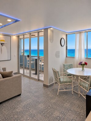 Een moderne kamer met een grote schuifglazen deur die leidt naar een balkon met uitzicht op de oceaan. Er staat een ronde eettafel met stoelen, een bank en een klok aan de muur.