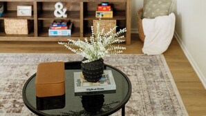 A living room with a round glass coffee table, a brown ottoman, a potted plant, and a magazine. A flat-screen TV is mounted on a wooden entertainment unit with storage boxes.