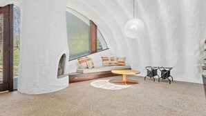 Une pièce chaleureuse avec un plafond voûté, une grande fenêtre, un coin salon avec des coussins, une table ronde et deux chaises.