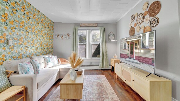 Ein Wohnzimmer mit floralem Wandpapier, einem weißen Sofa, einem Holztisch, einem Fernseher auf einem Holzständer und einem Fenster mit Vorhängen.