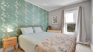 Ein Schlafzimmer mit blumigem Wandpapier, einem Bett mit gestreifter Bettwäsche, einem Nachttisch mit Lampe und einem Fenster mit Vorhängen.