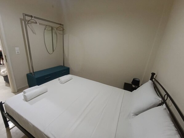 En pænt redt seng med hvide hør og puder, et lille natbord med en lampe, og et vægmonteret spejl med to knager.
