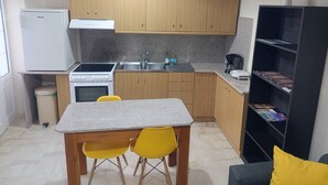 A compact kitchen with wooden cabinets, a gas stove, a sink, and a small refrigerator. There is a dining table with two chairs and a bookshelf with books. - Comfy studio near city center (Heraklion)