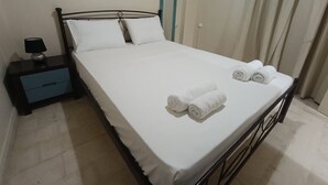 A neatly made bed with white linens and rolled towels, a bedside table with a lamp, and a blue drawer. - Comfy studio near city center (Heraklion)