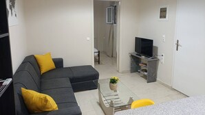 A living room with a grey sofa, yellow cushions, a glass coffee table, a TV on a stand, and a door leading to another room. - Comfy studio near city center (Heraklion)