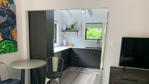 Een compact woongedeelte met een kitchenette, een eethoek met een ronde tafel en twee stoelen, een tv op een standaard en een boekenkast met boeken.