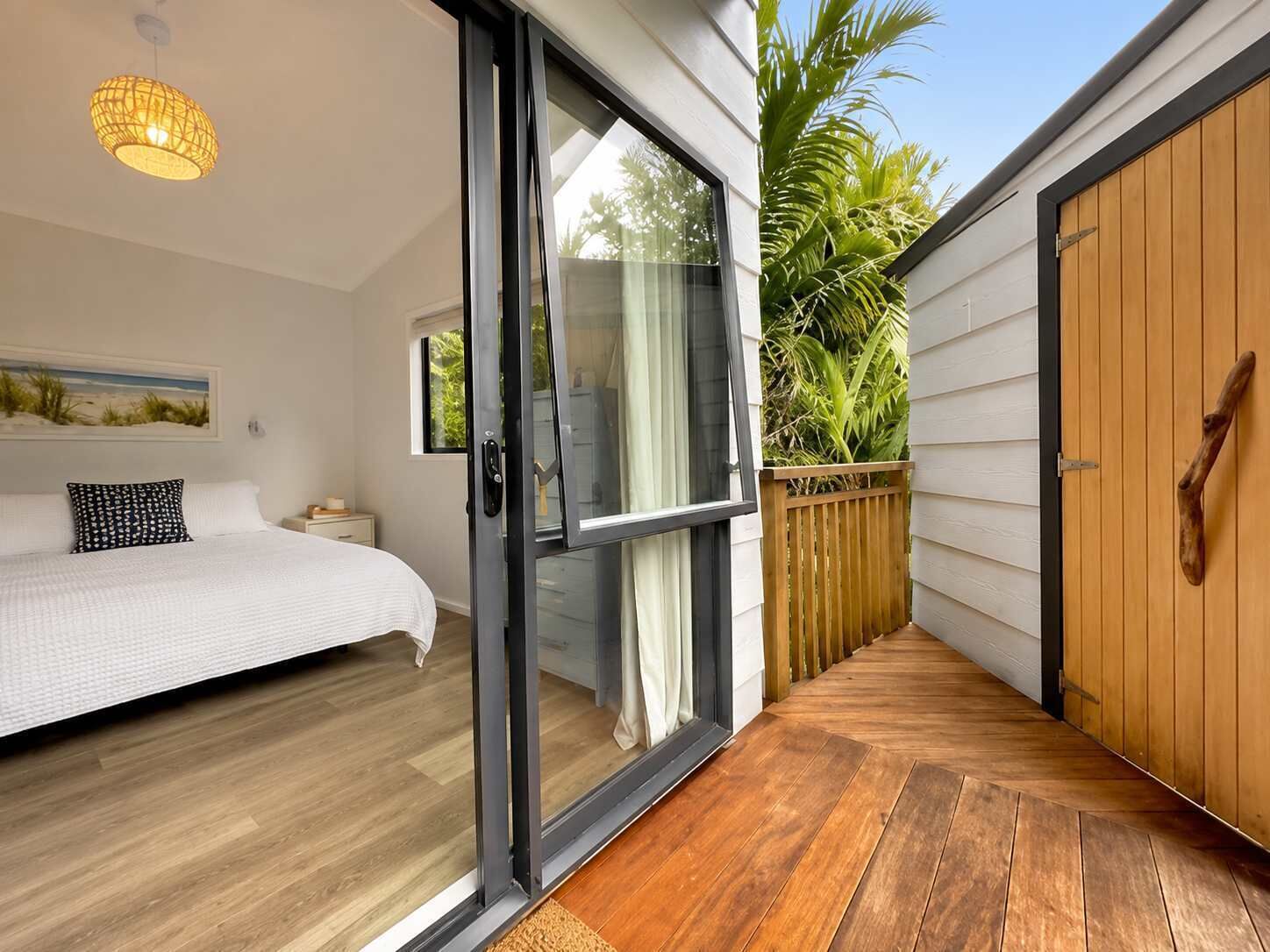 Ein Schlafzimmer mit einem Bett, einer Glasschiebetür, die auf eine Holzterrasse führt, und Blick auf eine Palme.