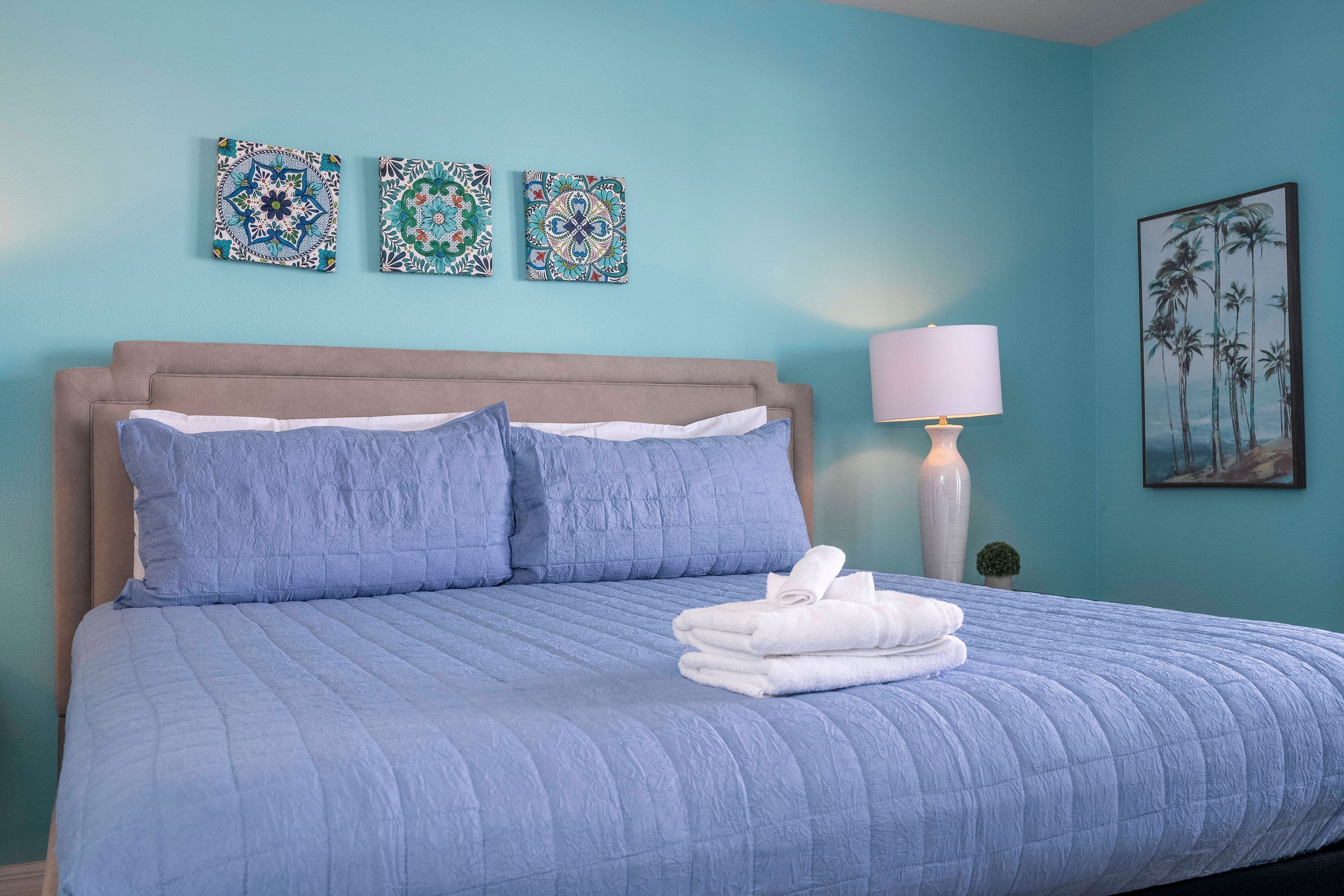 A bedroom with a bed, pillows, a folded towel, a lamp, and wall art.