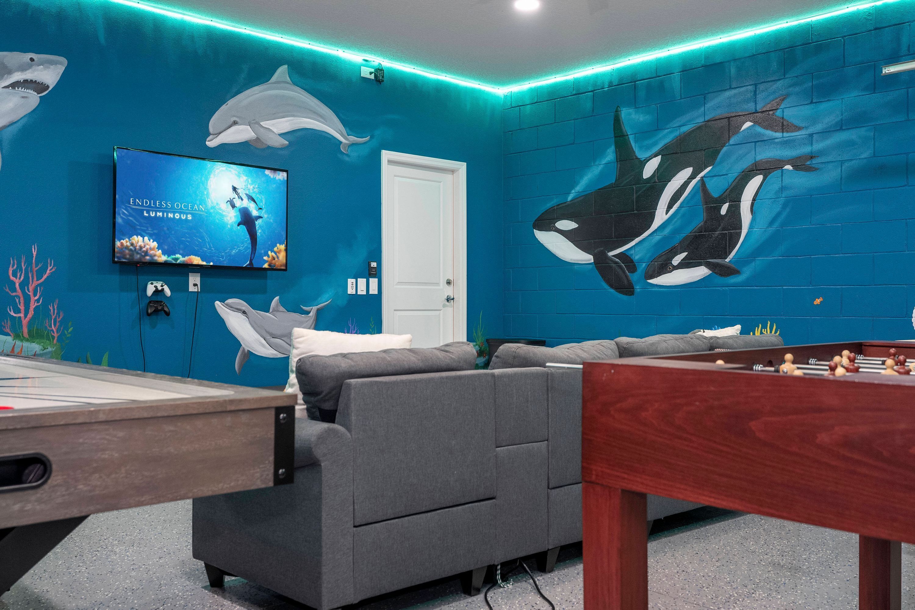 Une pièce avec une table de billard, un canapé gris, un téléviseur diffusant l’image de « Endless Ocean Luminescent », et une fresque représentant des orques.