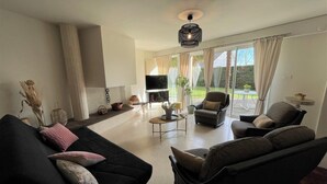 A modern living room with a black sofa, two armchairs, a coffee table, and a TV. There is a large window with curtains, a potted plant, and a hanging light fixture. - Lou Quillé - UZEIN - Gîte (UZEIN)