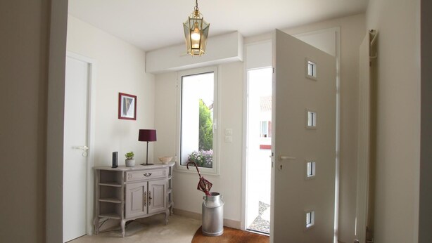 A hallway with a white door, a small cabinet with a lamp, a window with a view of greenery, and a trash can. - Lou Quillé - UZEIN - Gîte (UZEIN)