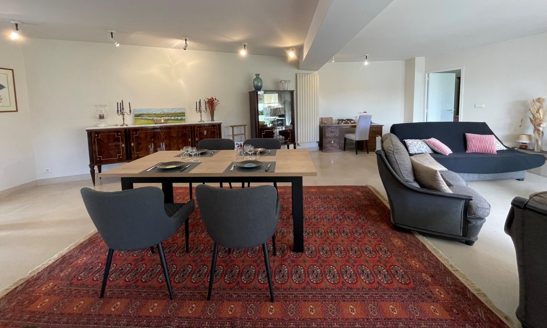 Uma sala de estar moderna com um sofá cinza escuro, uma área de jantar com mesa e cadeiras de madeira e um tapete estampado.