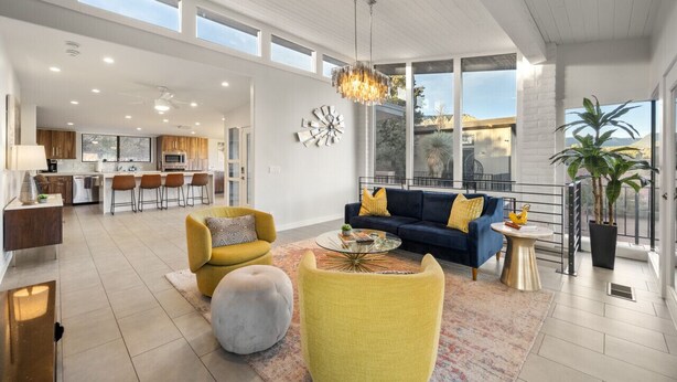 A spacious living area with a blue sofa, yellow armchairs, and a round coffee table. A large chandelier hangs from the ceiling, and there is a view of the kitchen and dining area in the background. - Endless RedRock Views Modern Uptown Home w/Spa/Gym (Sedona)