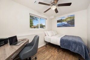 A bedroom with a bed, a desk with a laptop, a chair, a ceiling fan, a window, and a painting. - Rmnp Retreat - Group-friendly Home Near Estes Park (Loveland)
