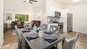 Un coin repas avec une table dressée pour quatre personnes, une cuisine équipée d’appareils modernes et un salon avec un canapé et un tableau.