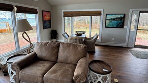 A spacious living room with a brown leather sofa, a coffee table, and wicker chairs. Large windows offer a view of the outdoors.