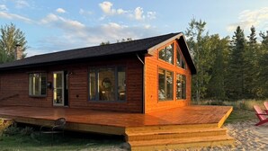 A wooden cabin with a deck, large windows, and a sandy beach in the background.