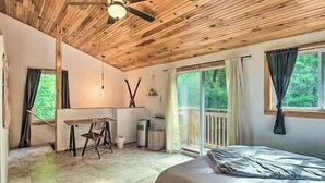 A bedroom with a bed, a desk, and a chair. There is a window with curtains and a view of greenery outside.