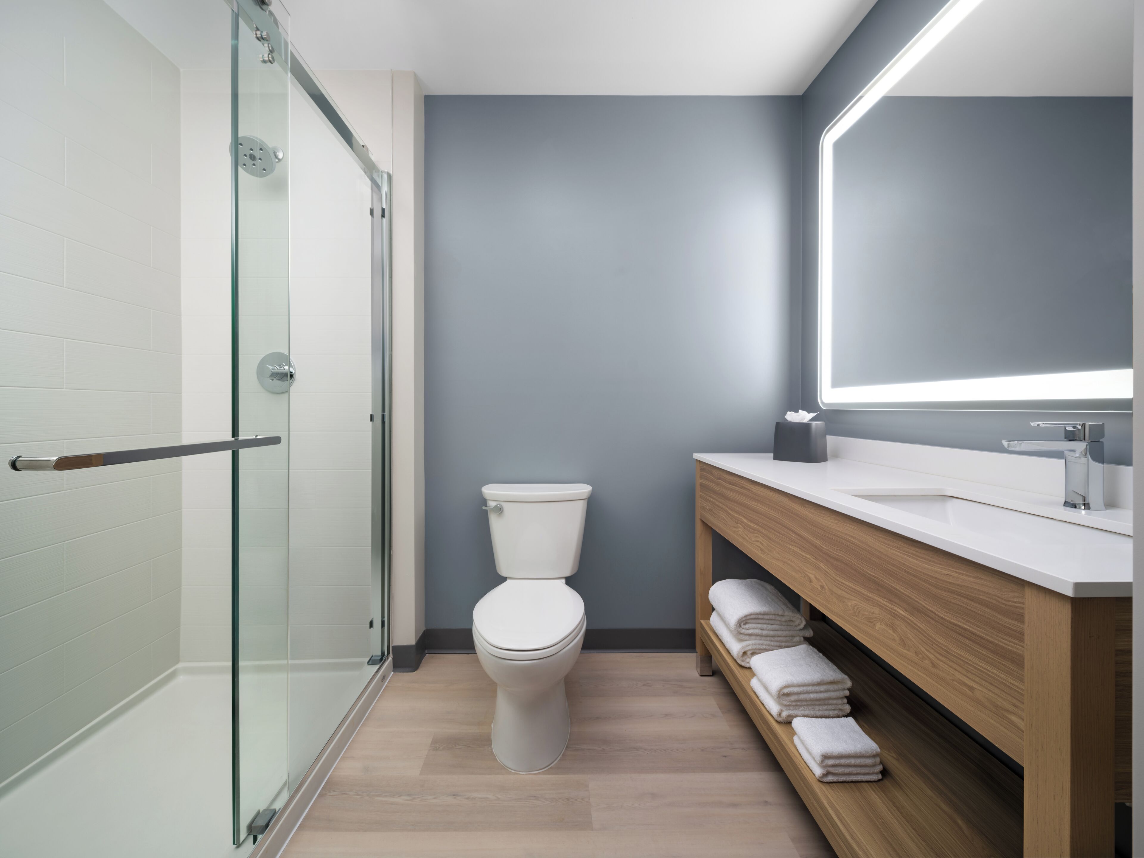 A modern bathroom with a glass shower enclosure, a white toilet, a wooden vanity with a mirror, and neatly folded towels.