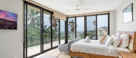 Une chambre avec un grand lit, une chaise et une vue sur les palmiers et les maisons, visible à travers de grandes fenêtres.