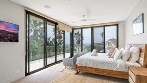 A bedroom with a large bed, a chair, and a view of palm trees and houses through large windows. - Clifftop Luxury With Pool ~ Ultimate outdoor experience with a private pool (Kiama)